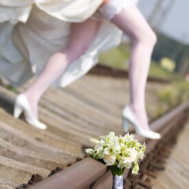 mały bukiet ślubny z białych storczyków i róż oparty na torach. w tle panna młoda stojąca na torach, pokazująca podwiązkę