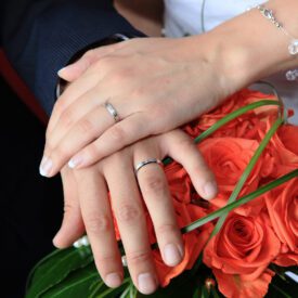 dłonie państwa młodych z obrączkami oparte o bukiet ślubny z pomarańczowych róż